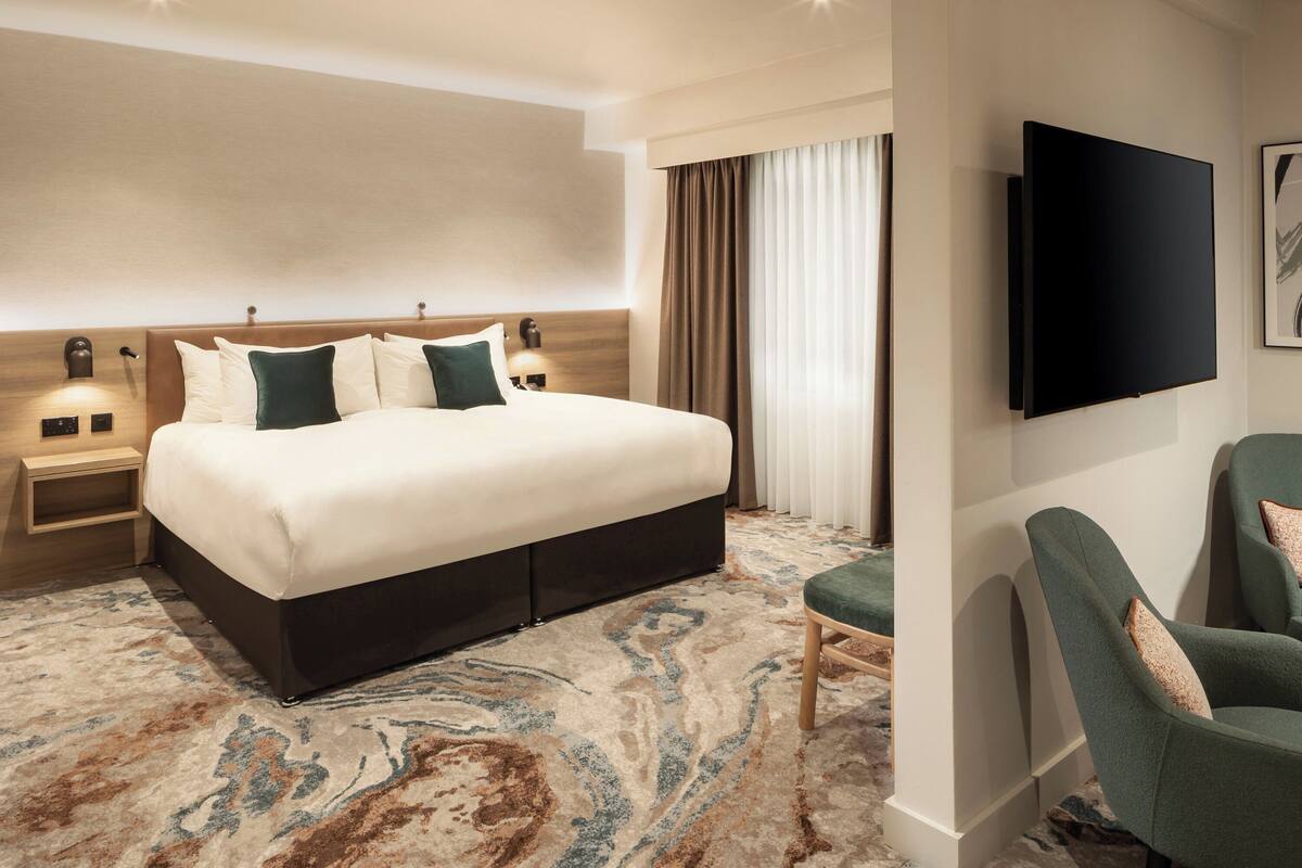 A hotel room with a large bed, a flat-screen TV, a green armchair, and a patterned carpet.