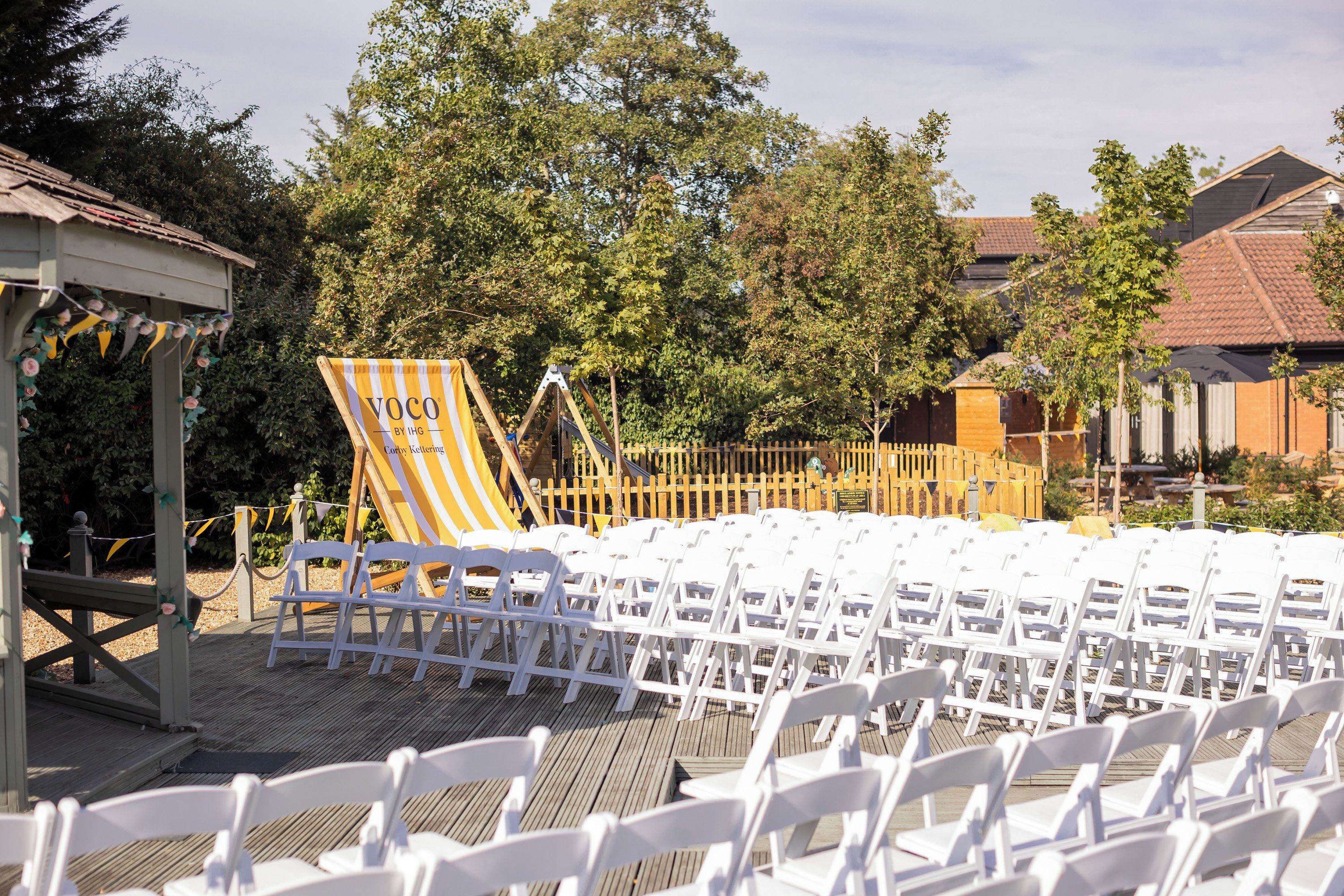 A deck with white chairs arranged for an event, a striped tent, and a wooden fence in the background.