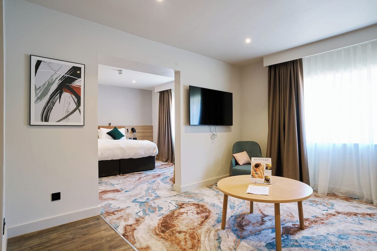 A hotel room with a bed, a TV, a chair, and a table with a book.
