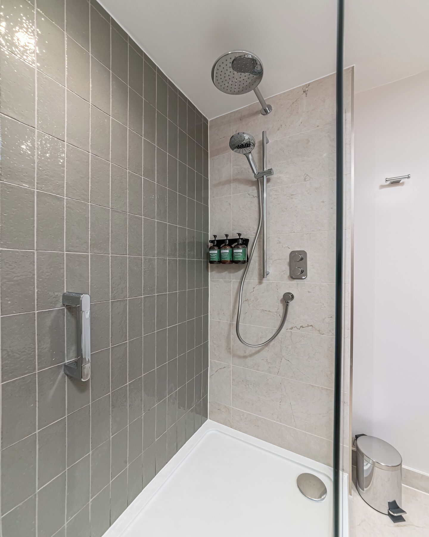 Et moderne bruseområde med brusehoved i hånden, et fastmonteret brusehoved og et brusearmatur i hånden. Tre flasker shampoo og balsam står på en hylde.