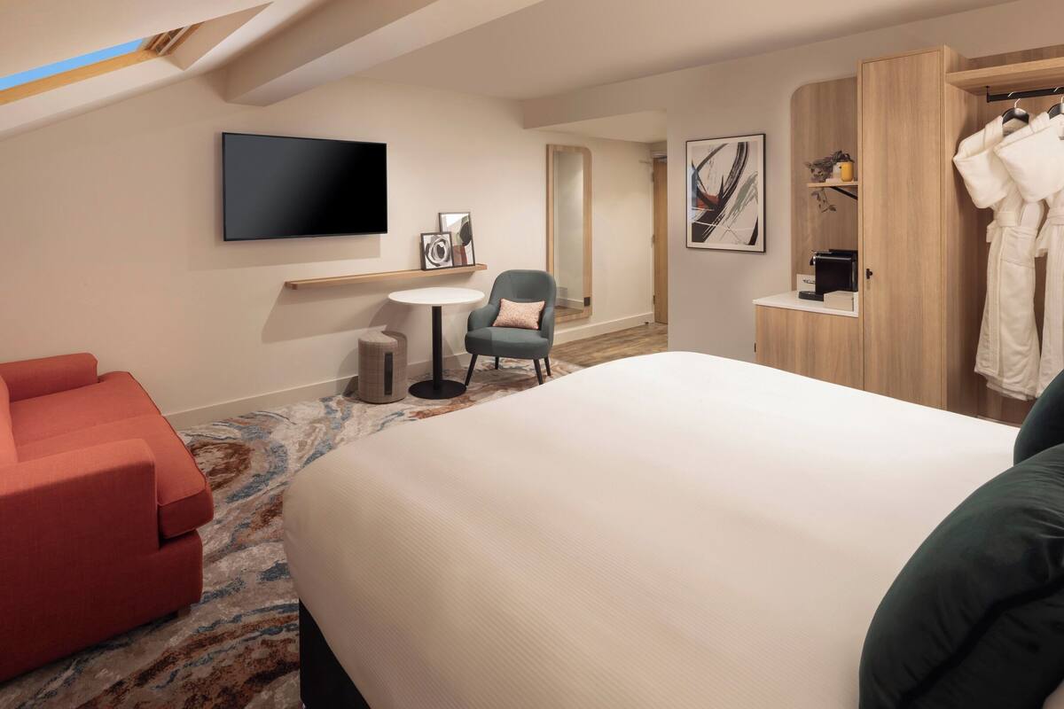 A hotel room with a bed, a red sofa, a small table, a chair, a TV, a mirror, and a wardrobe.