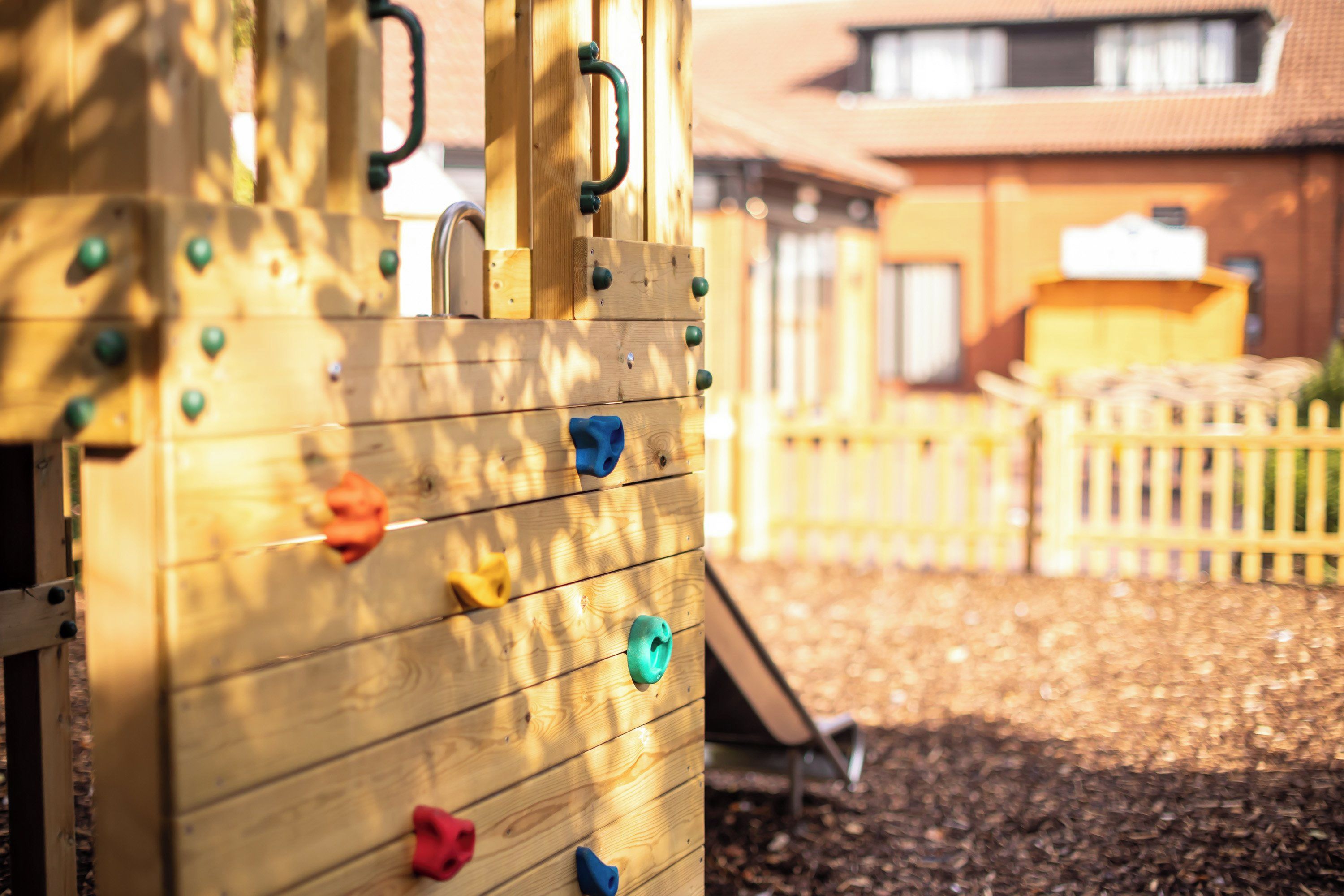 A wooden climbing structure with various handholds and a slide, set against a backdrop of a building and a fence.