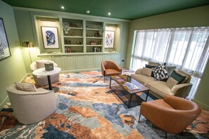 Un salón con un sofá, sillones, una mesa de centro y una alfombra estampada. Se ve una estantería con varios objetos y cuadros colgados en la pared.