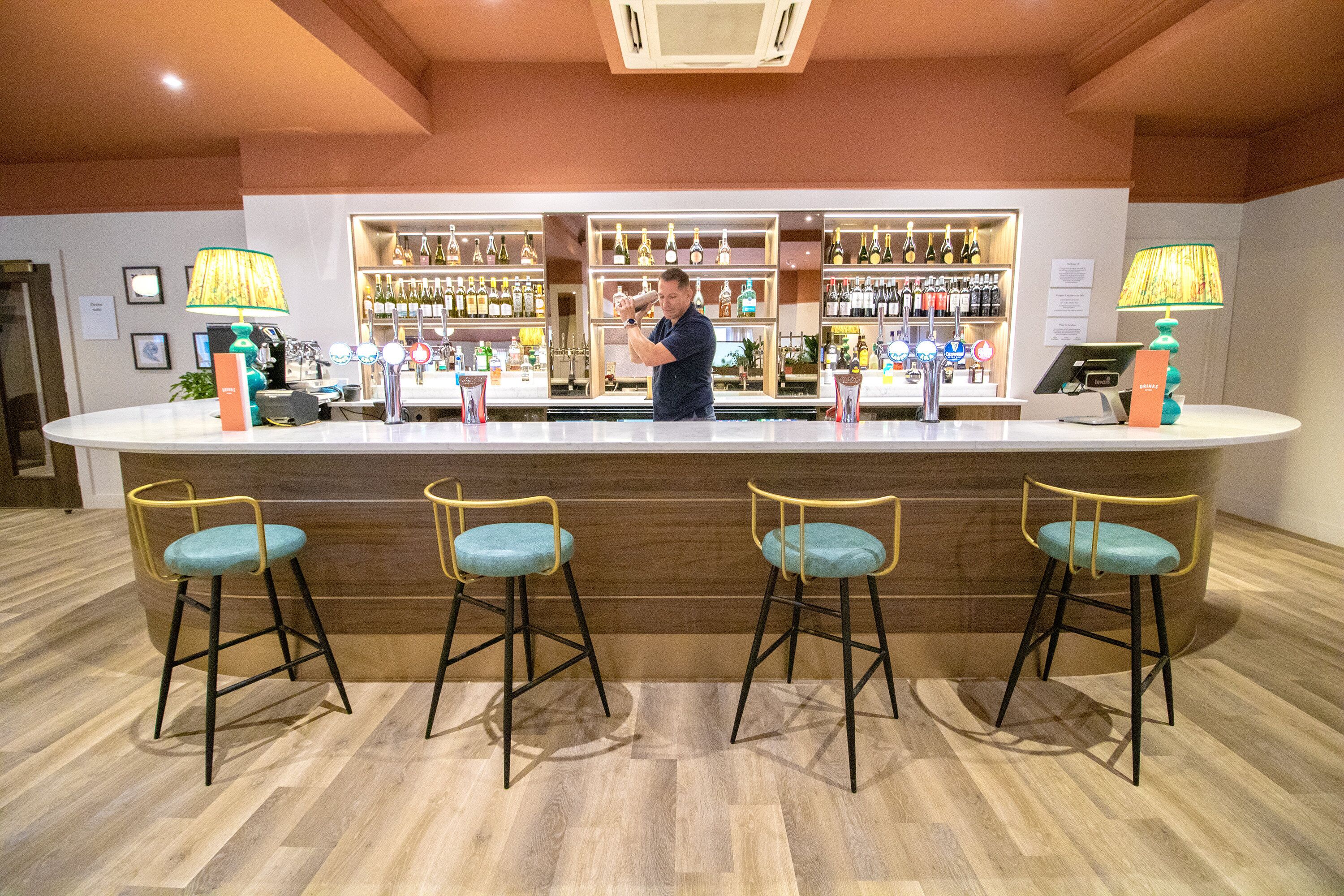 A bar area with a bartender, wooden bar counter, and three bar stools with teal cushions.