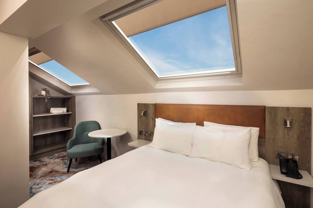 A hotel room with a skylight, a bed, a desk, a chair, and a small shelf.