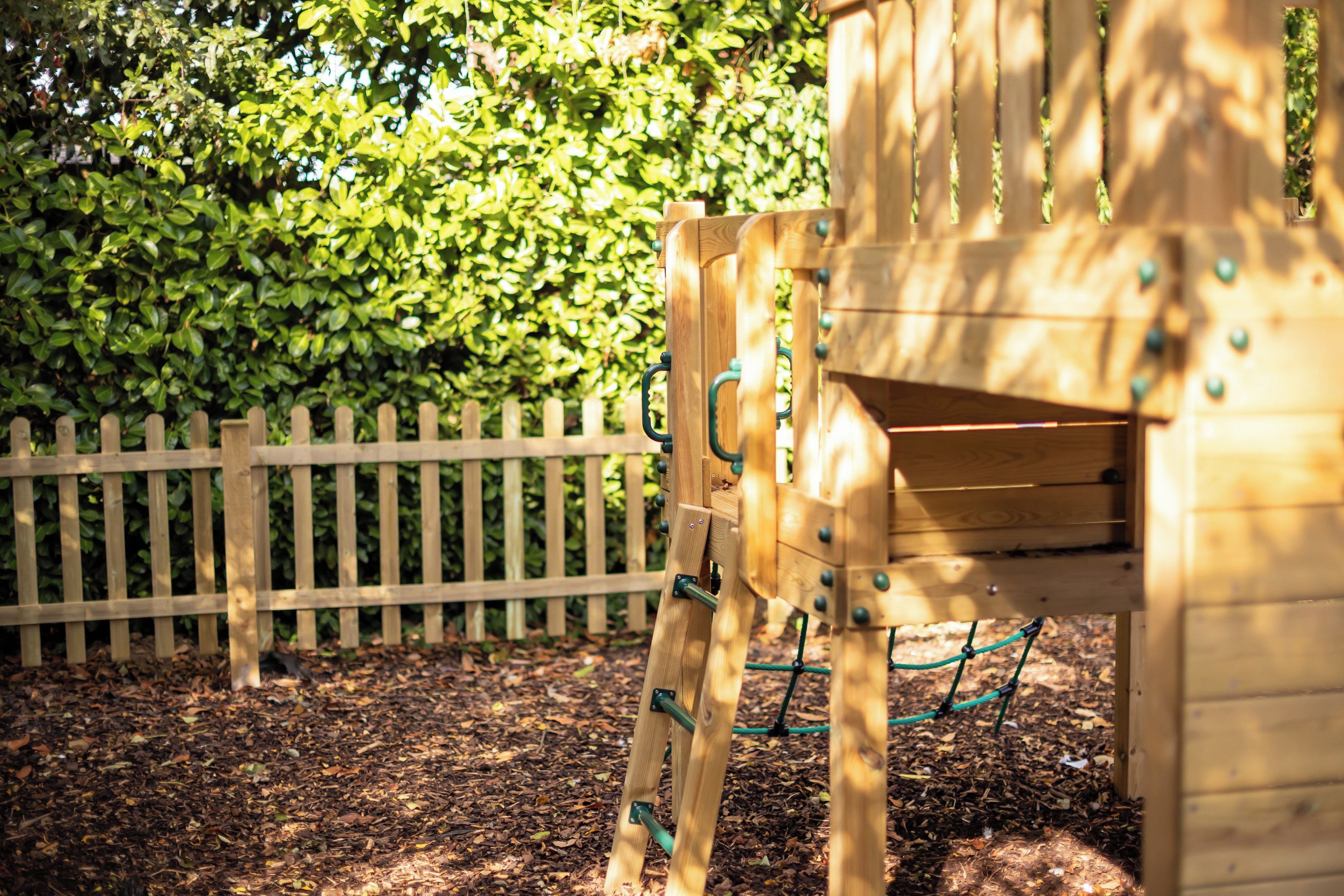 A wooden play structure with a ladder and a slide, surrounded by a wooden fence and a hedge.