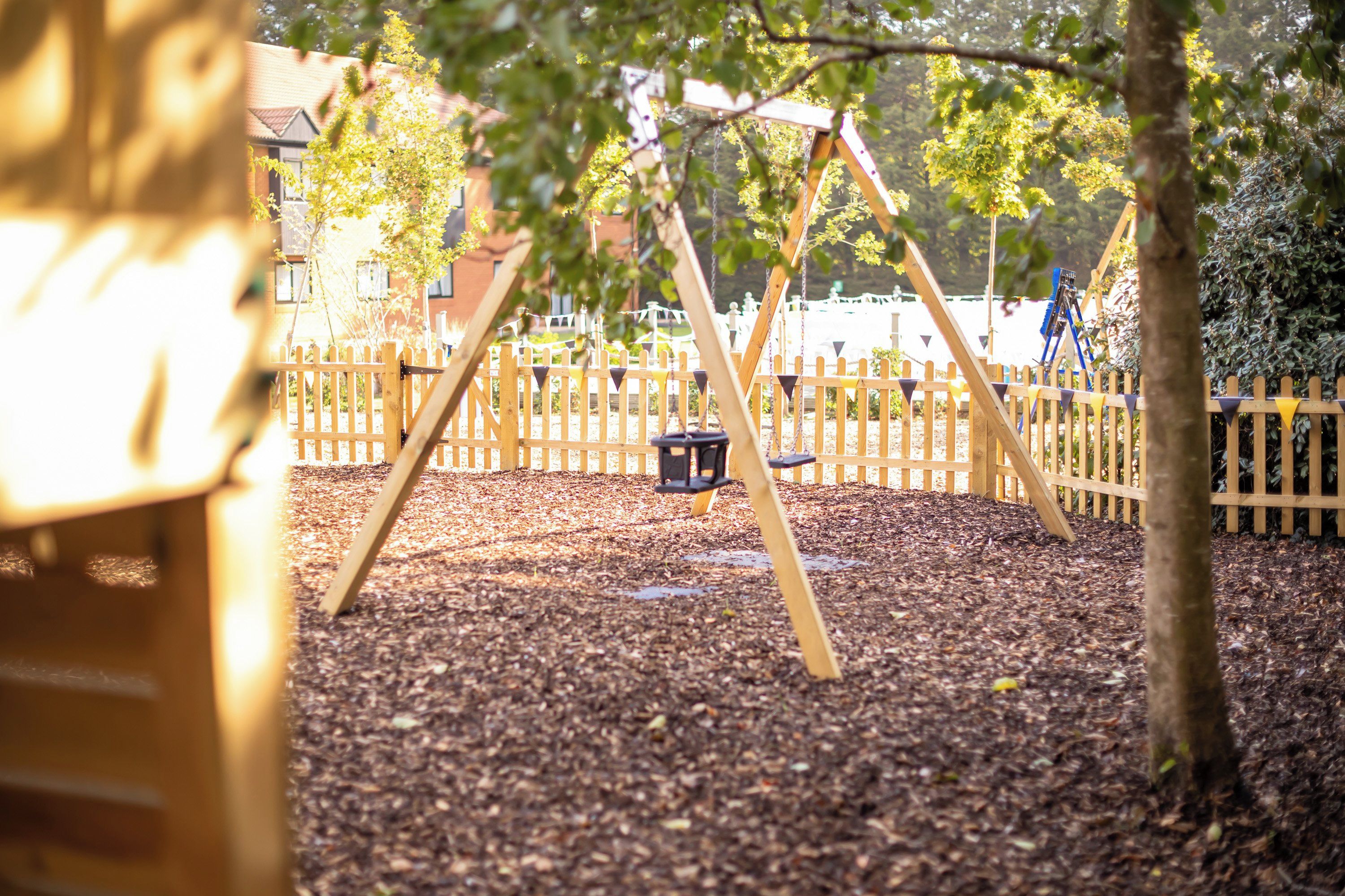 A playground with a swing set and a slide, surrounded by a wooden fence and trees.