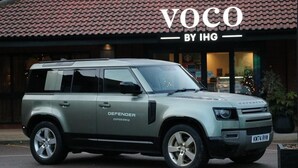 Een zilveren Defender SUV geparkeerd voor een gebouw met een pannendak en een bord waarop 'VOCO BY IHG' staat.