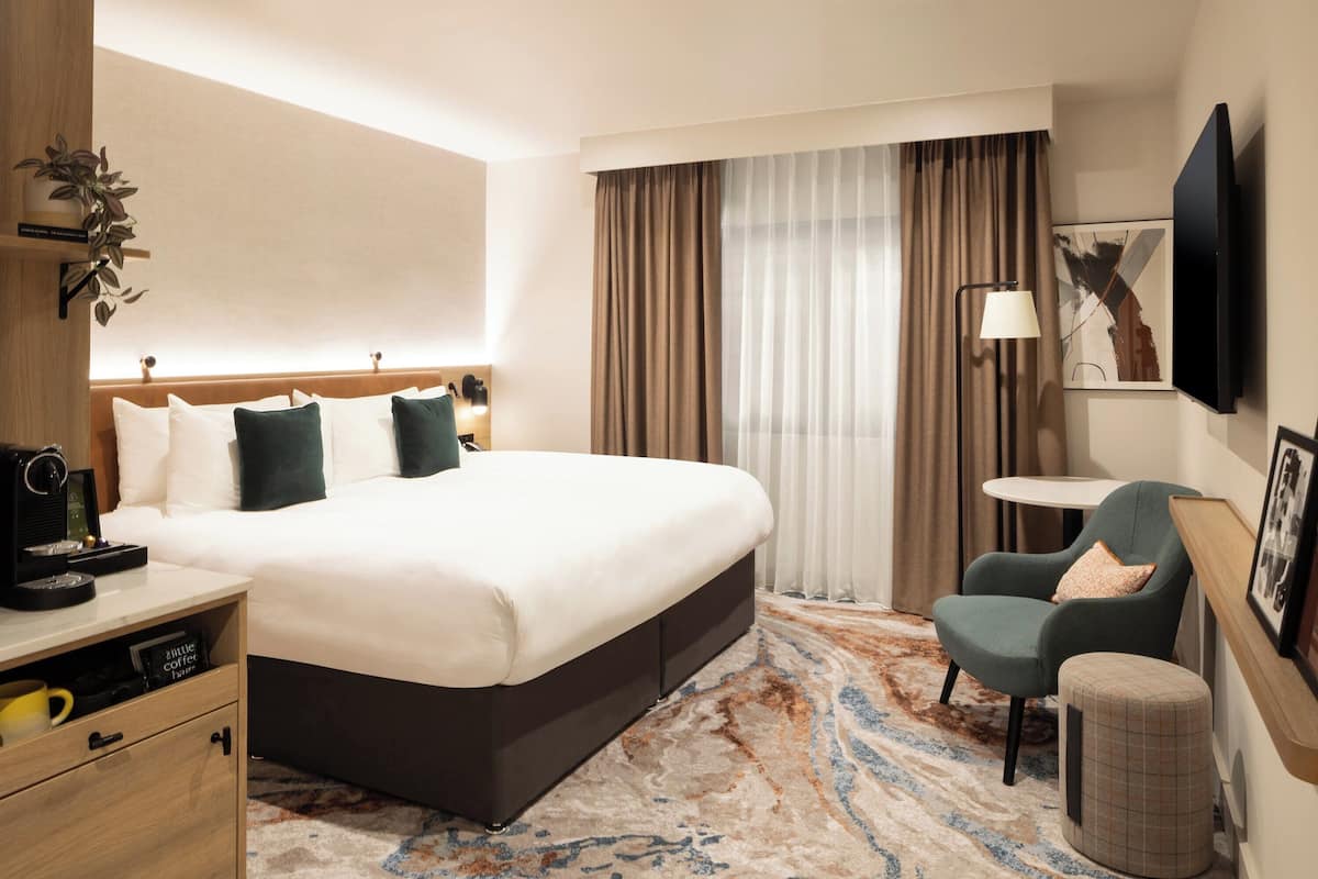 A hotel room with a large bed, a desk with a coffee machine, a chair, a small table, a TV, and a large window with curtains.