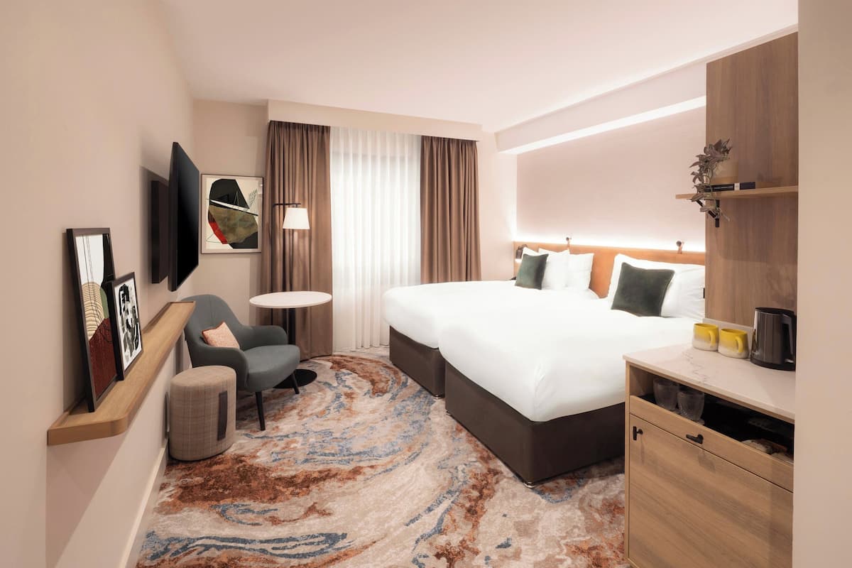 A hotel room with a bed, a chair, a small table, a TV, and a desk with a lamp.