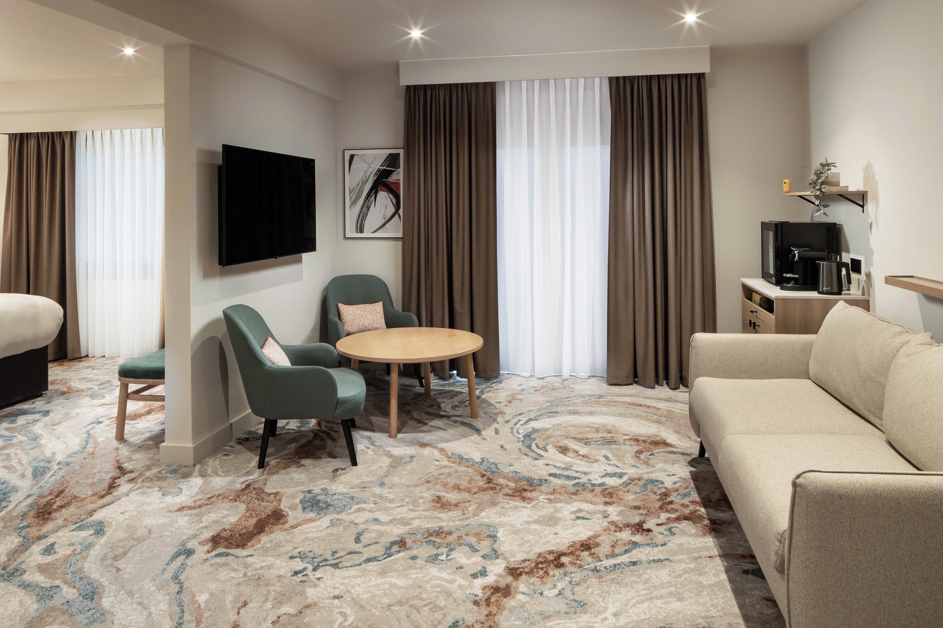 A hotel room with a sofa, two armchairs, a small round table, a flat-screen TV, a coffee machine, and a large window with curtains.