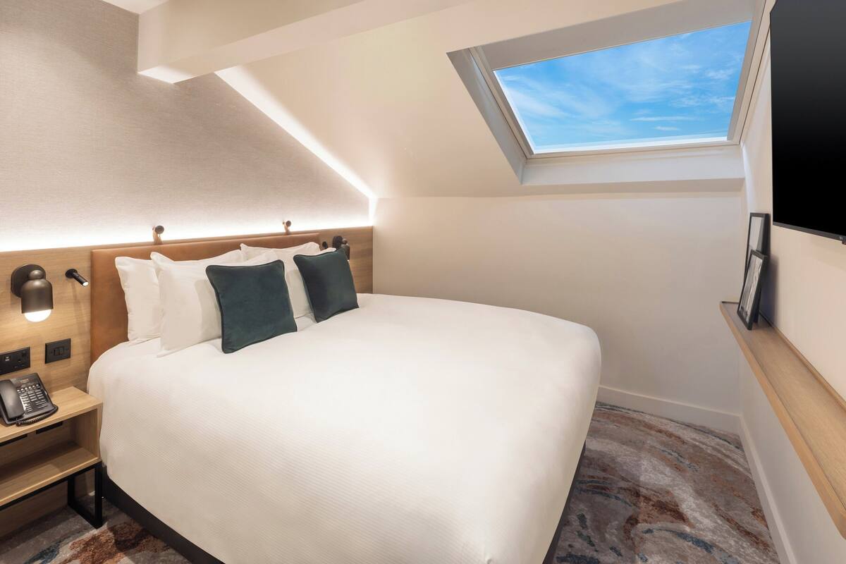 A hotel room with a large bed, a skylight, a bedside telephone, and a wooden headboard.