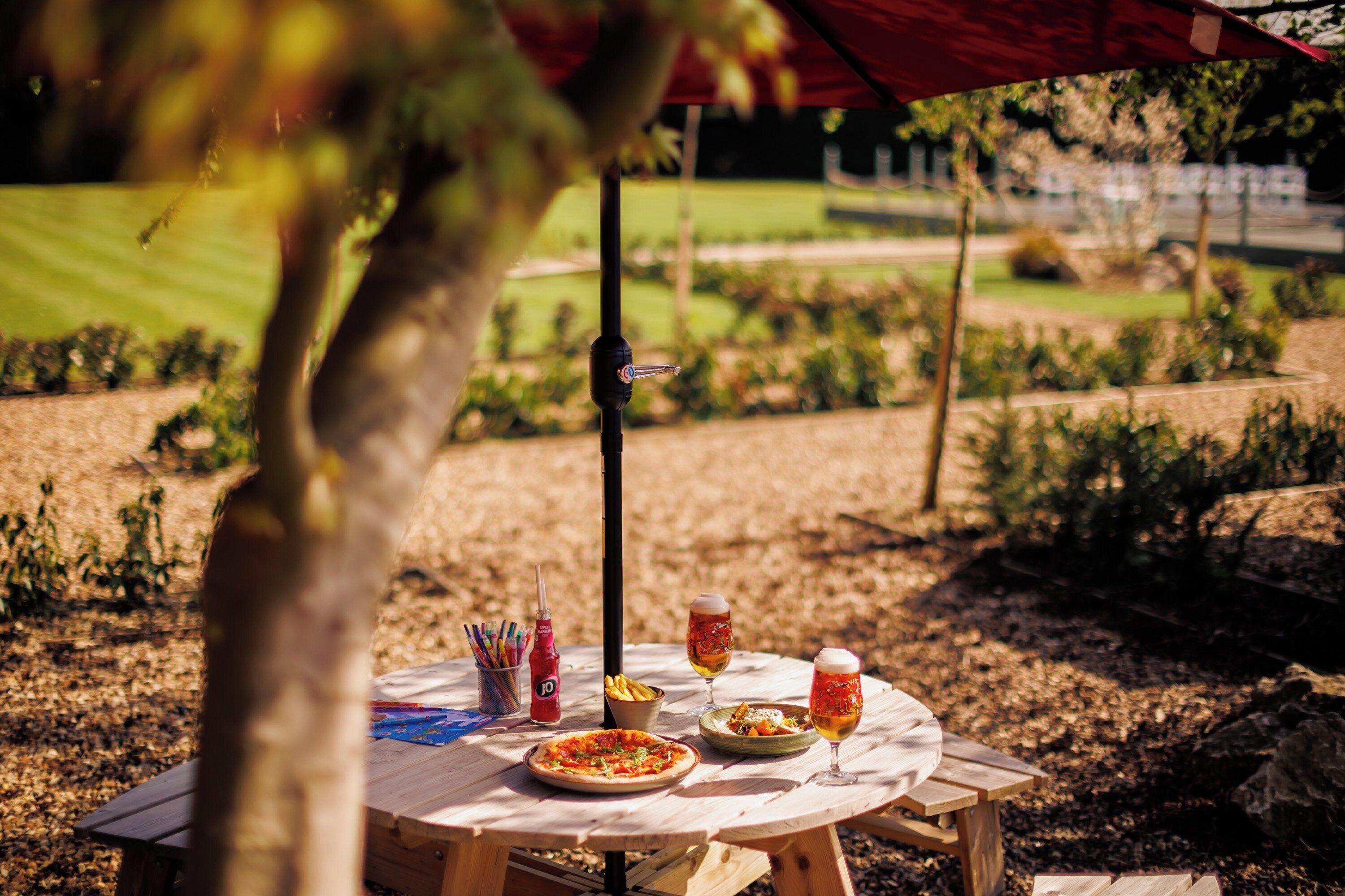 A round wooden table with a pizza, two glasses of beer, a bottle of ketchup, and a bowl of chips.