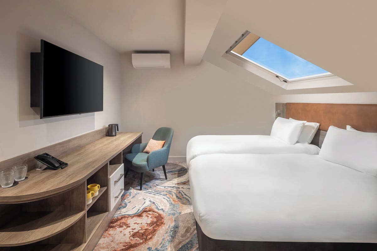 A hotel room with a flat-screen TV, a desk with a telephone, two beds, a chair, and a patterned rug.