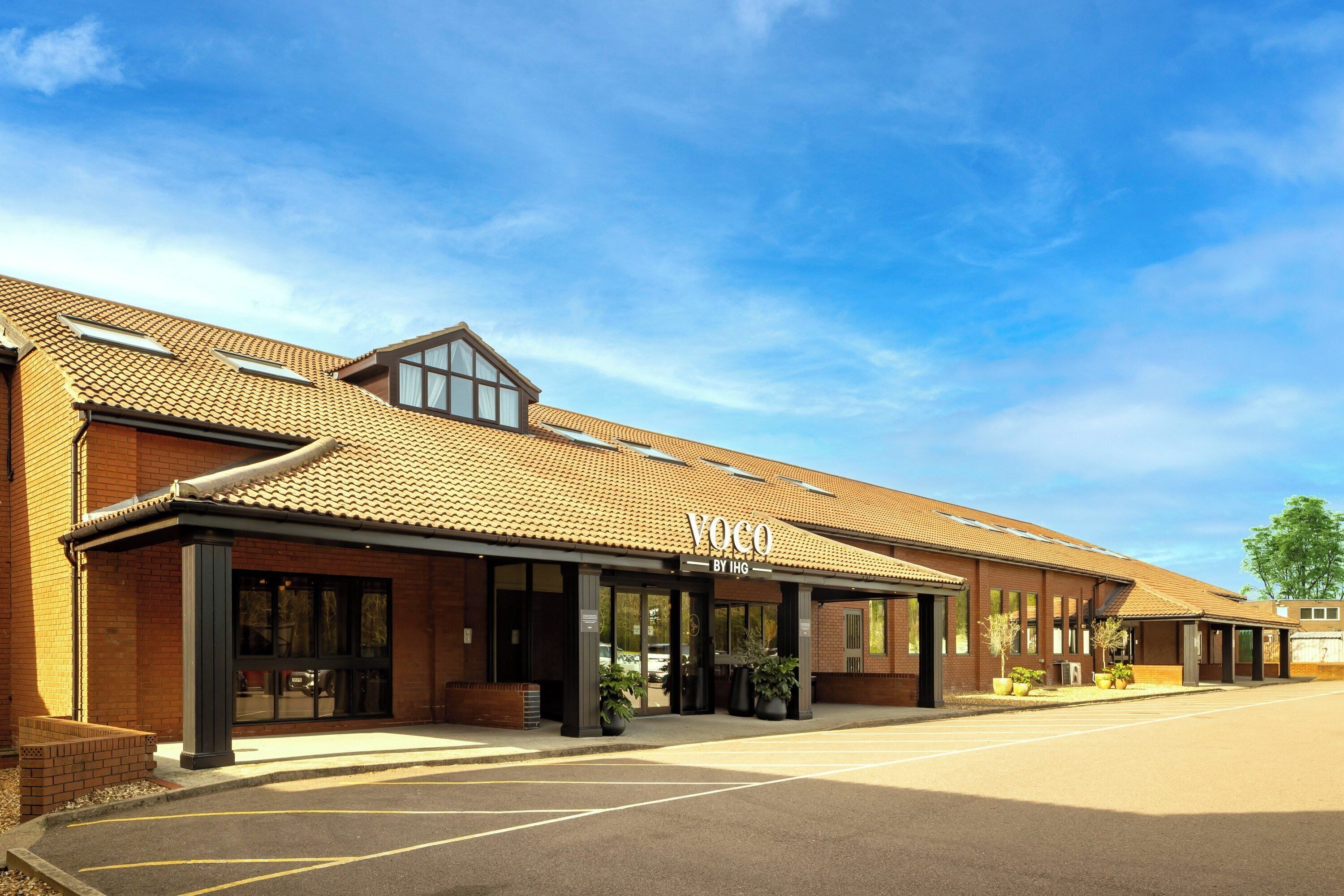 A building with a tiled roof, a sign reading 'Voco By IHG,' and a parking lot.