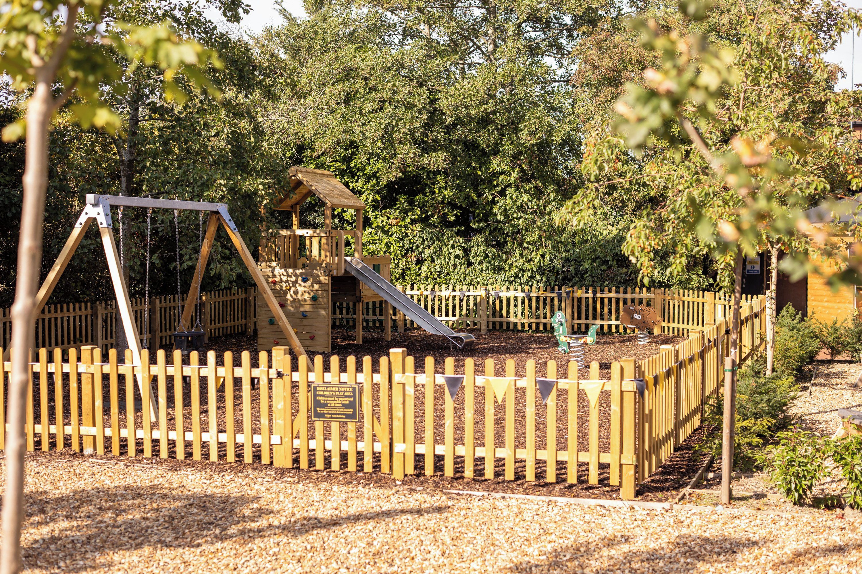 Ein Spielplatz mit Holzschaukel, Rutsche und Klettergerüsten, umgeben von einem Holenzaun und Bäumen.
