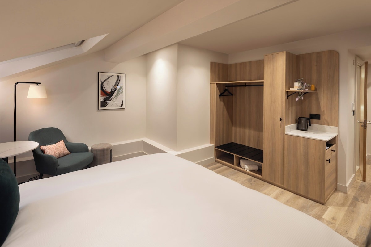 A modern hotel room with a bed, a seating area with a chair, a desk, a wardrobe, and a small shelf with a plant.