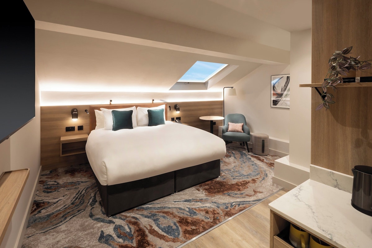 Un dormitorio moderno con tragaluces, una cama grande, mesitas de noche, una silla y una pequeña mesa redonda.