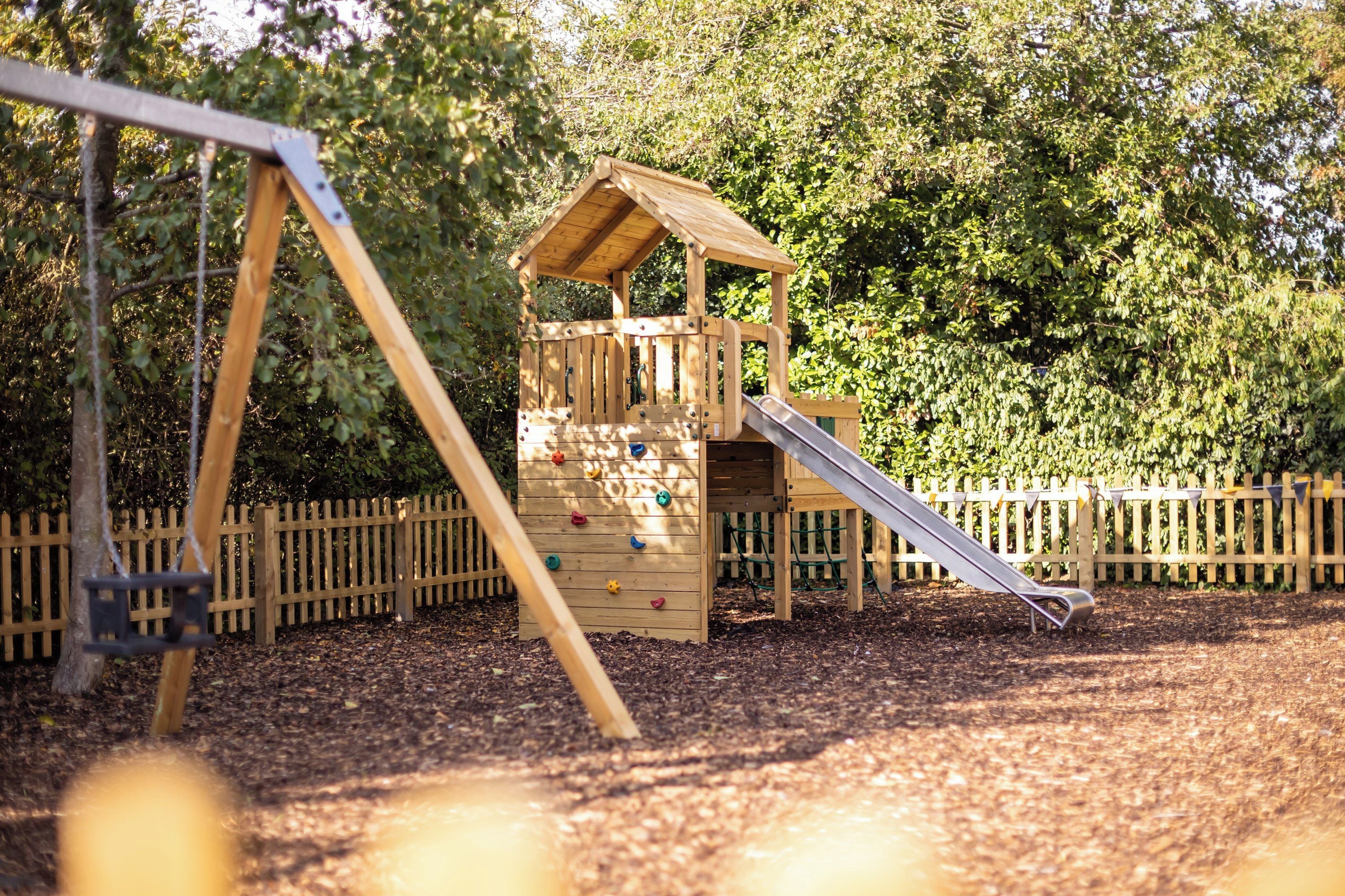 A wooden play structure with a slide and climbing wall, surrounded by a wooden fence and trees.
