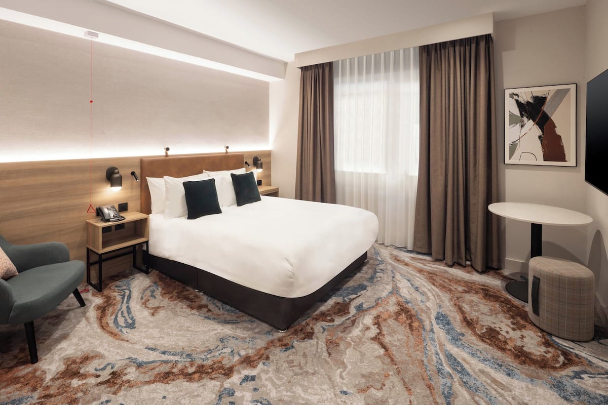 A hotel room with a large bed, bedside tables, a chair, a desk, a TV, and a patterned carpet.