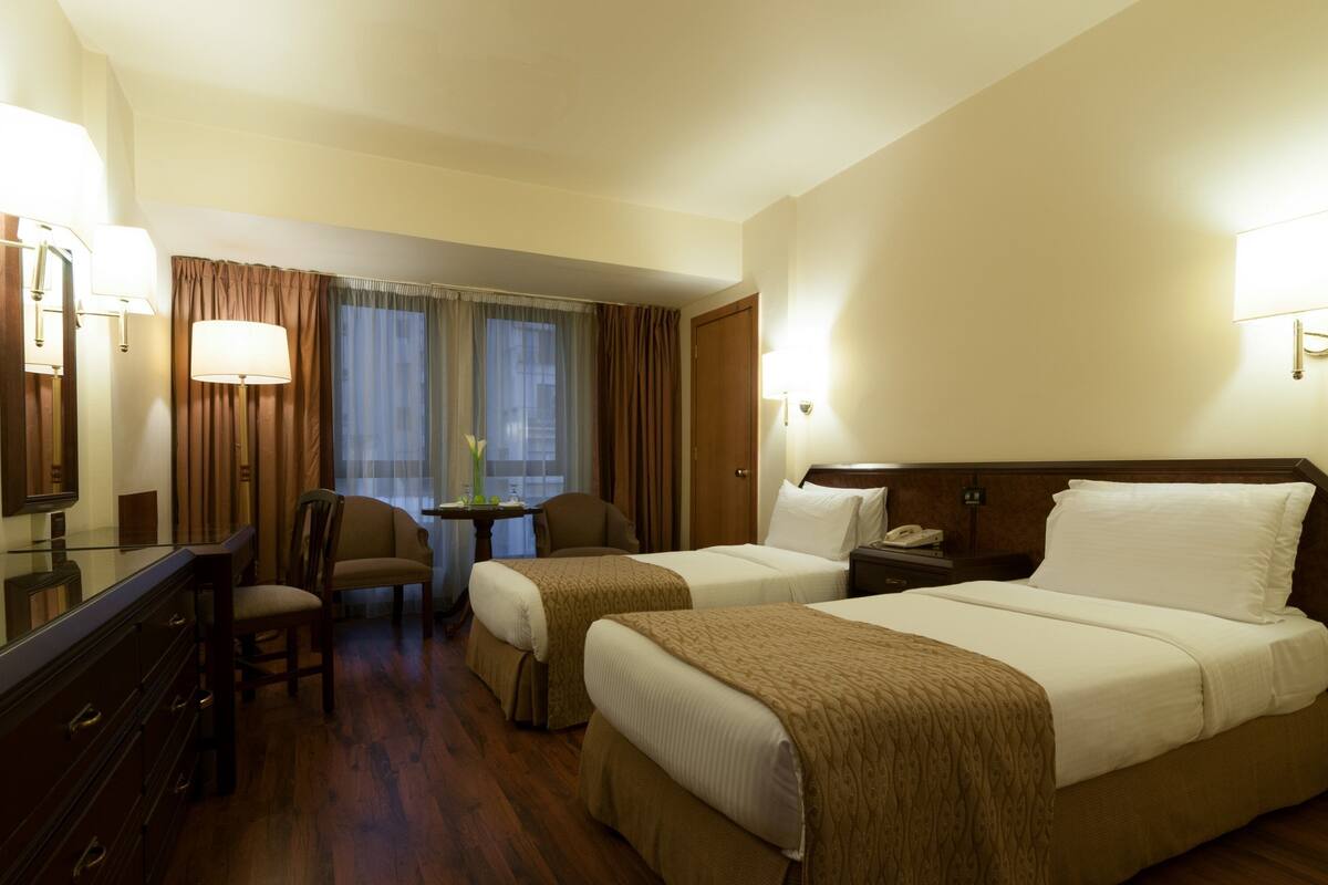A hotel room with two beds, a desk, a chair, and a small table with a lamp.