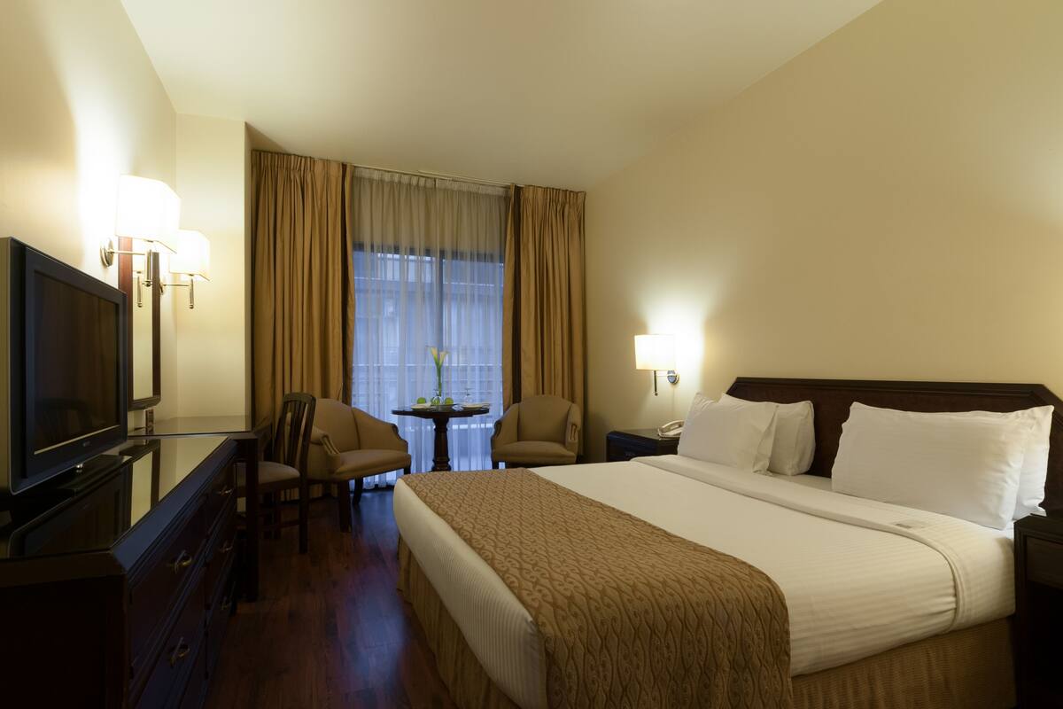 A hotel room with a bed, a TV, a desk with a chair, a small table with a vase, and a window with curtains.