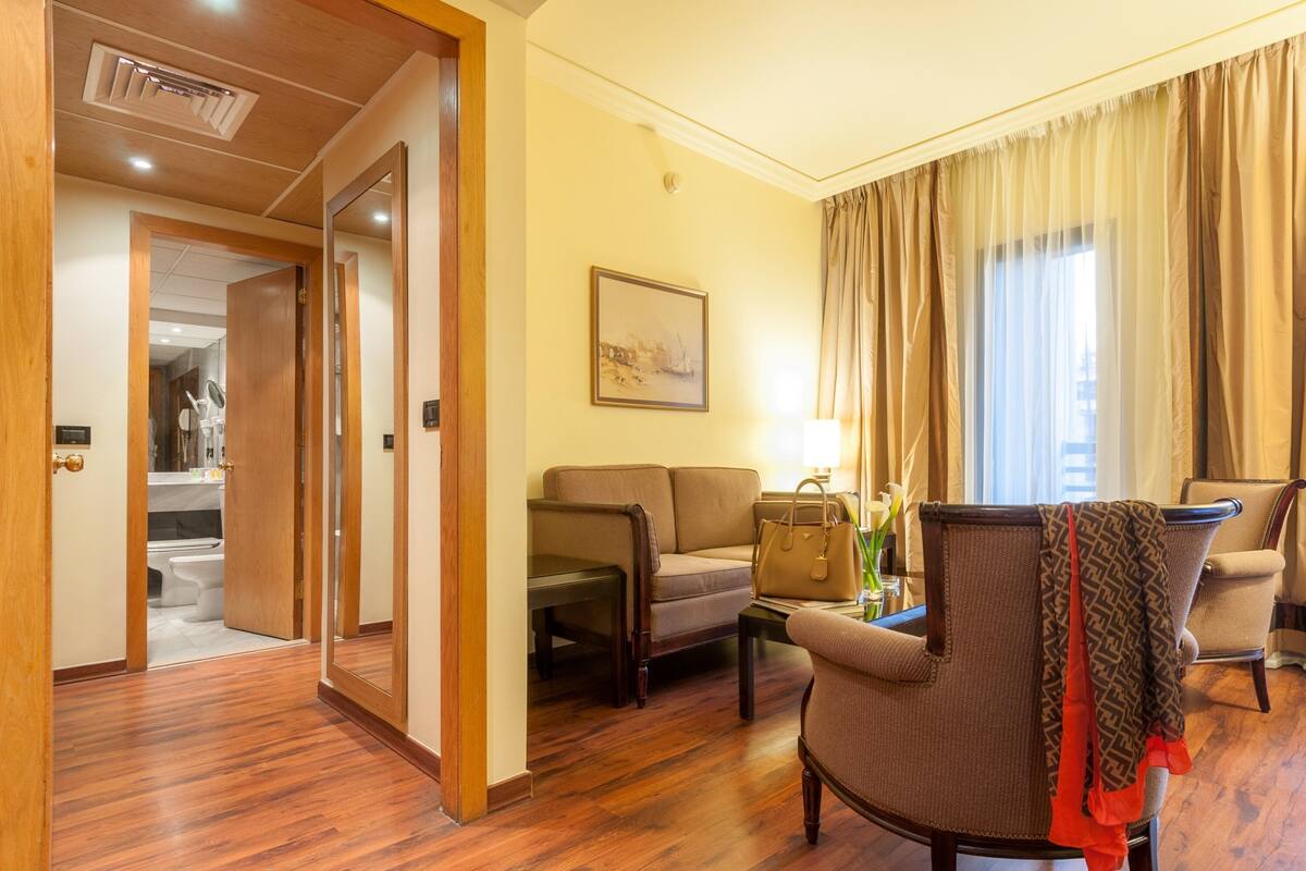 A hotel room with a wooden floor, a sofa, a chair, a table with a lamp, and a painting on the wall.