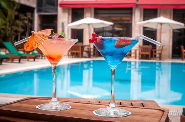 Two cocktails on a wooden tray by a poolside with umbrellas and lounge chairs in the background.
