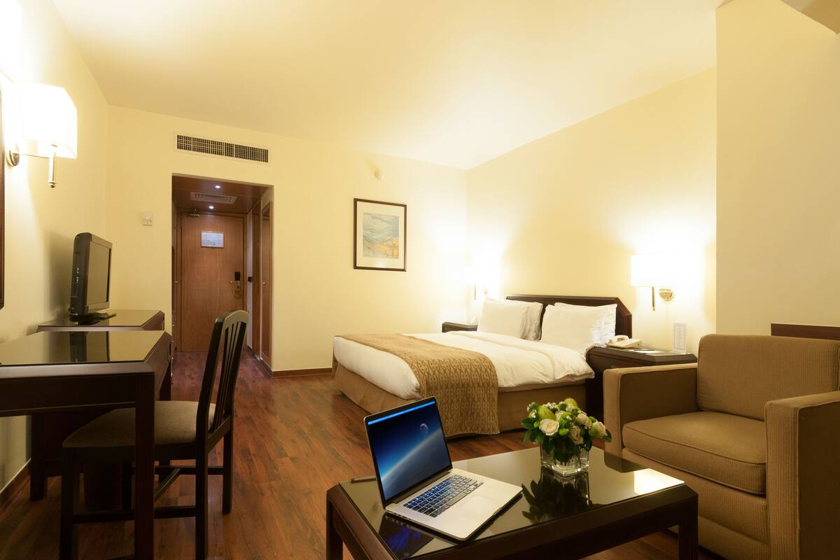 A hotel room with a bed, a desk with a laptop, a chair, a sofa, and a small table with a vase of flowers.