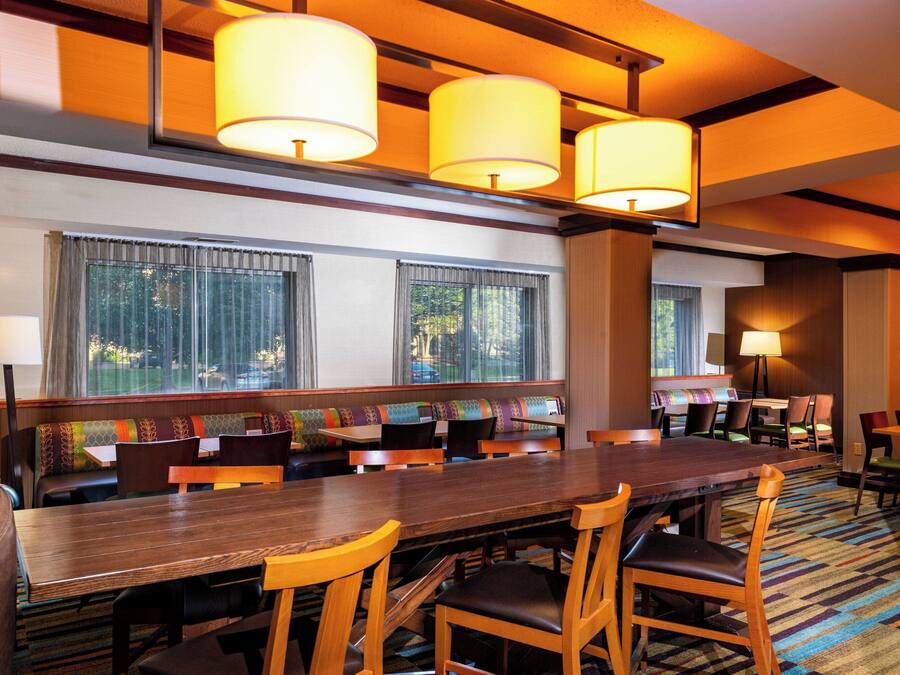 A hotel dining area with a long wooden table, multiple chairs, and a bench with colorful cushions. There are large windows with curtains, and the room has a warm, yellow-lit ceiling.