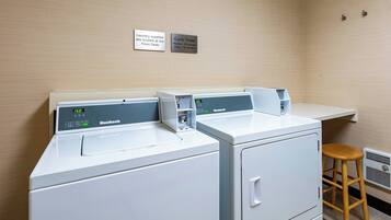 A laundry area with two front-loading machines, a wooden stool, and signs indicating laundry supplies and cycle times.
