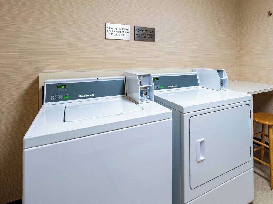 A laundry area with two front-loading machines, a wooden stool, and signs indicating laundry supplies and cycle times.