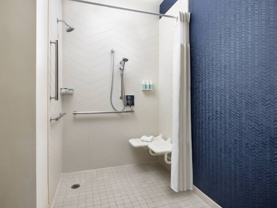 A shower area with a white tiled floor, a showerhead, a handheld shower, and a small shelf with toiletries.