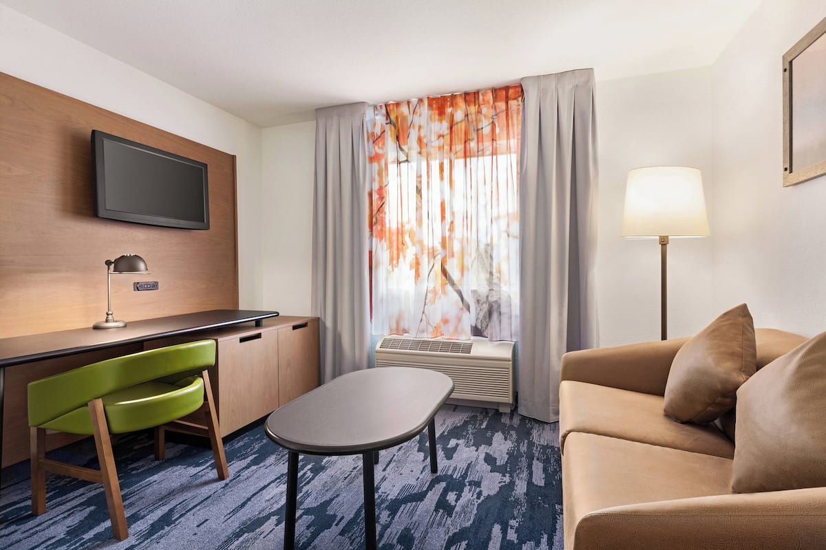 A hotel room with a desk, a green chair, a sofa, a TV mounted on the wall, a window with curtains, and a floor lamp.