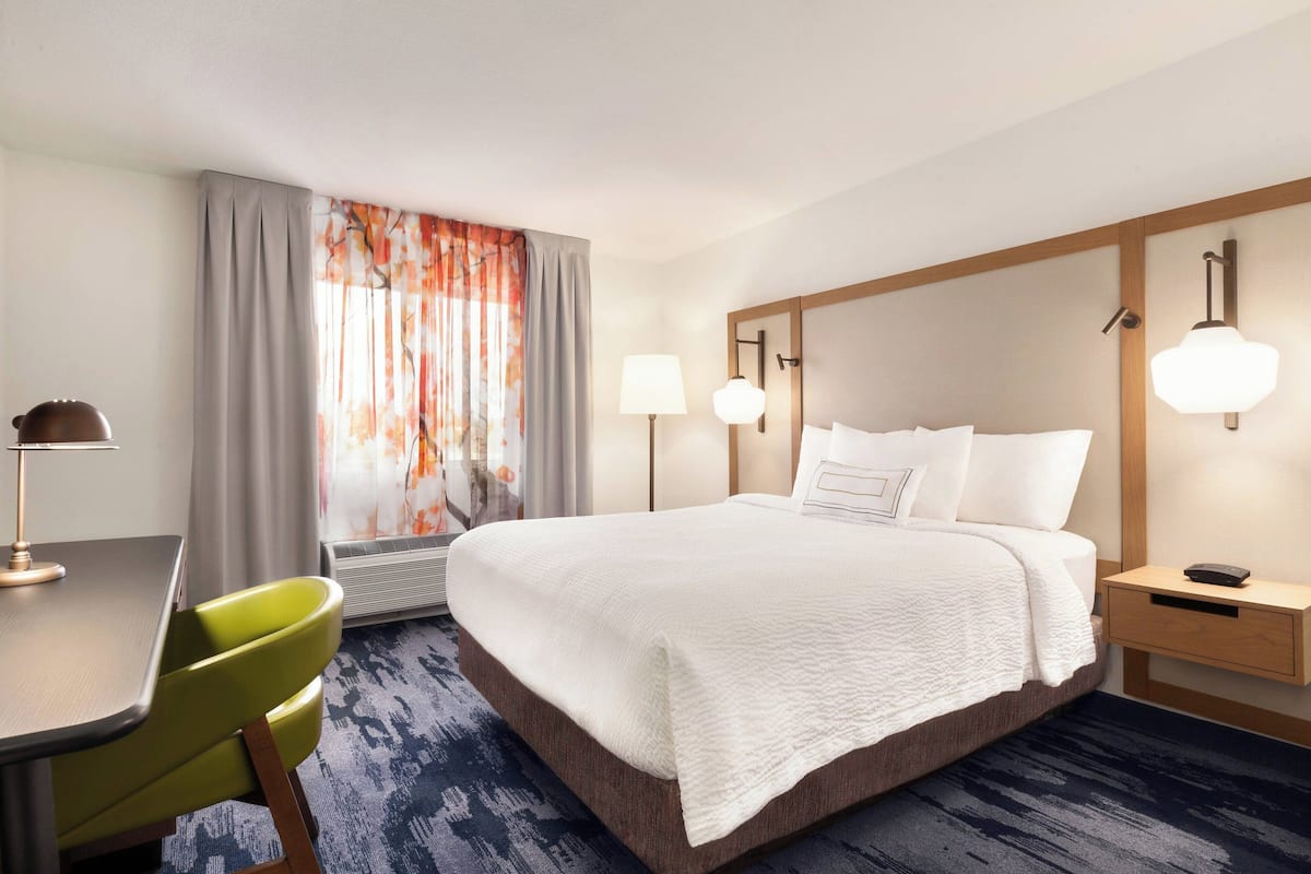 A hotel room with a bed, bedside table, lamp, chair, and a window with curtains.