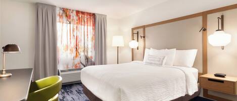 Quarto de hotel com cama, mesa de cabeceira, abajur, cadeira e janela com cortinas.