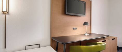 Habitación de hotel compacta con escritorio de madera, televisor montado en la pared, una silla verde y una mesita de noche.