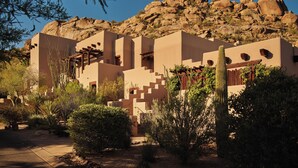 Un edificio de estilo desértico con varias habitaciones, rodeado de cactus y arbustos, y una montaña rocosa al fondo.