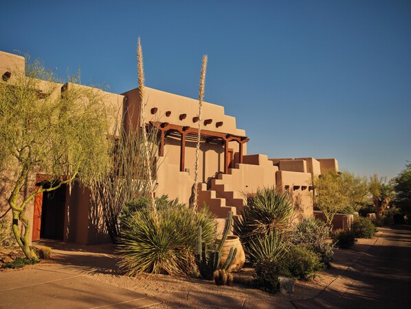 Traditional adobe-style building with a flat roof and covered walkway, surrounded by desert vegetation.