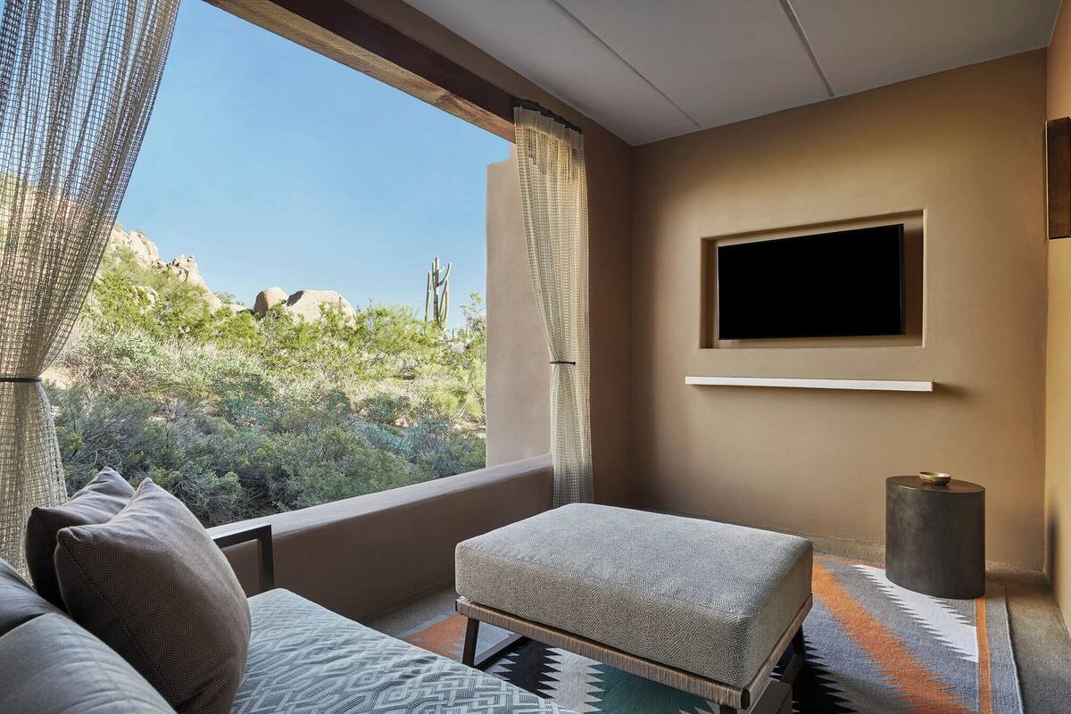 Een moderne kamer met een groot raam, een flatscreen televisie aan de muur, een klein voetenbankje en een kamerkleed met een patroon.