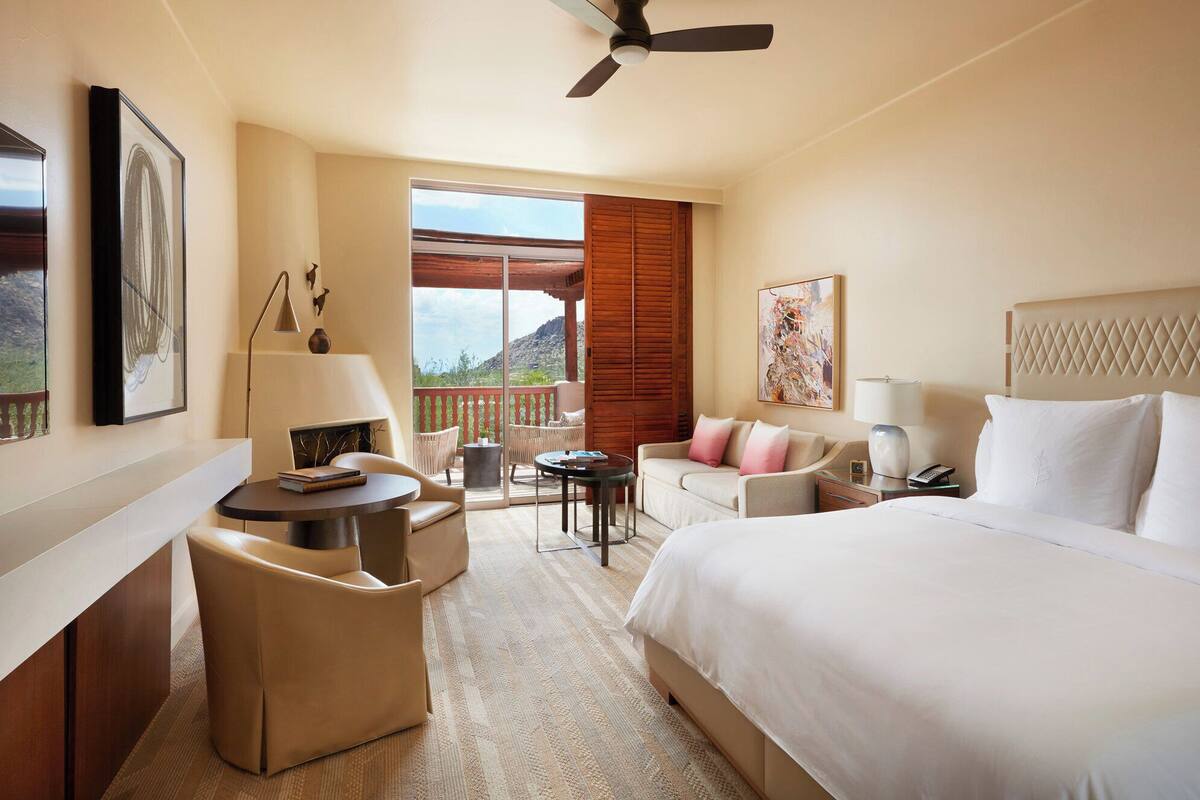 Een hotelkamer met een bed, een bureau met een stoel, een bank, een balkon met uitzicht op de bergen en een plafondventilator.