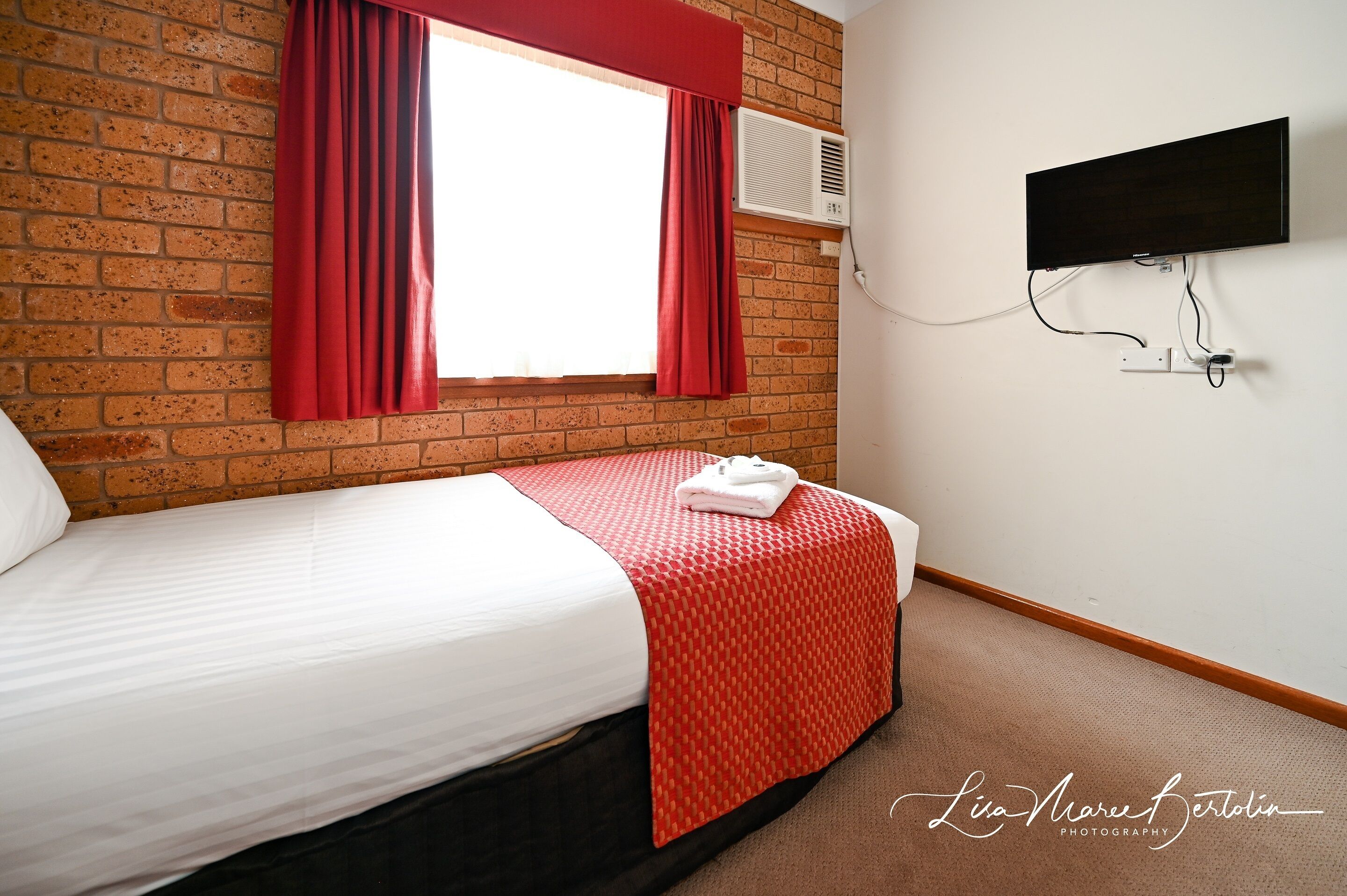 A hotel room with a brick wall, a bed with a white and red bedspread, a flat-screen TV mounted on the wall, and a window with red curtains.