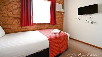 A hotel room with a brick wall, a bed with a white and red bedspread, a flat-screen TV mounted on the wall, and a window with red curtains.
