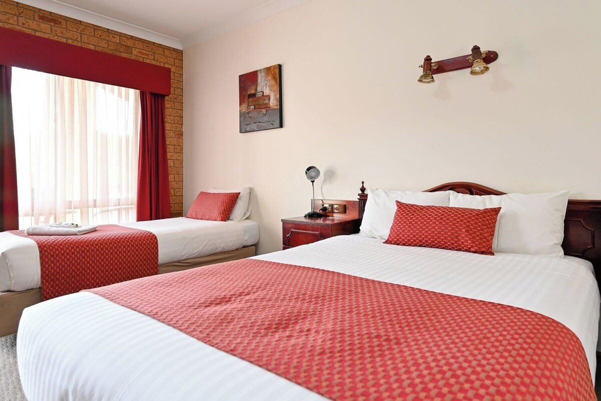 A hotel room with two beds, a wooden headboard, a nightstand, a lamp, and a painting on the wall.