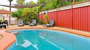 A swimming pool with a red fence, lounge chairs, and a shaded area.