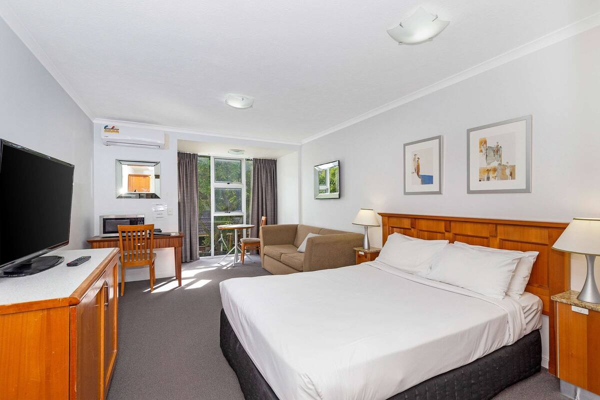 Ein Hotelzimmer mit Bett, Schreibtisch mit Fernseher, Sessel, Sofa, Lampe und gerahmten Bildern an der Wand.