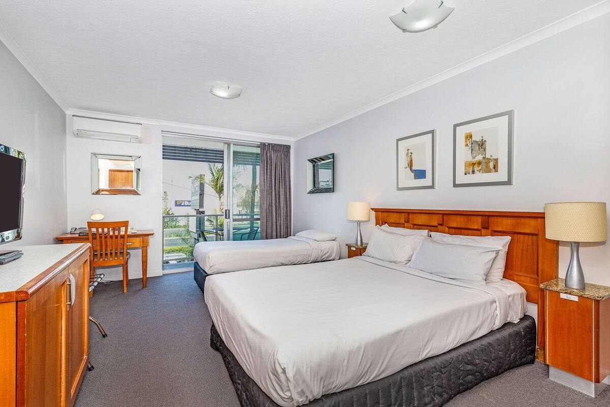Ein Hotelzimmer mit zwei Betten, einem Schreibtisch mit Stuhl, einem Fernseher und einem Balkon mit Blick auf Palmen.