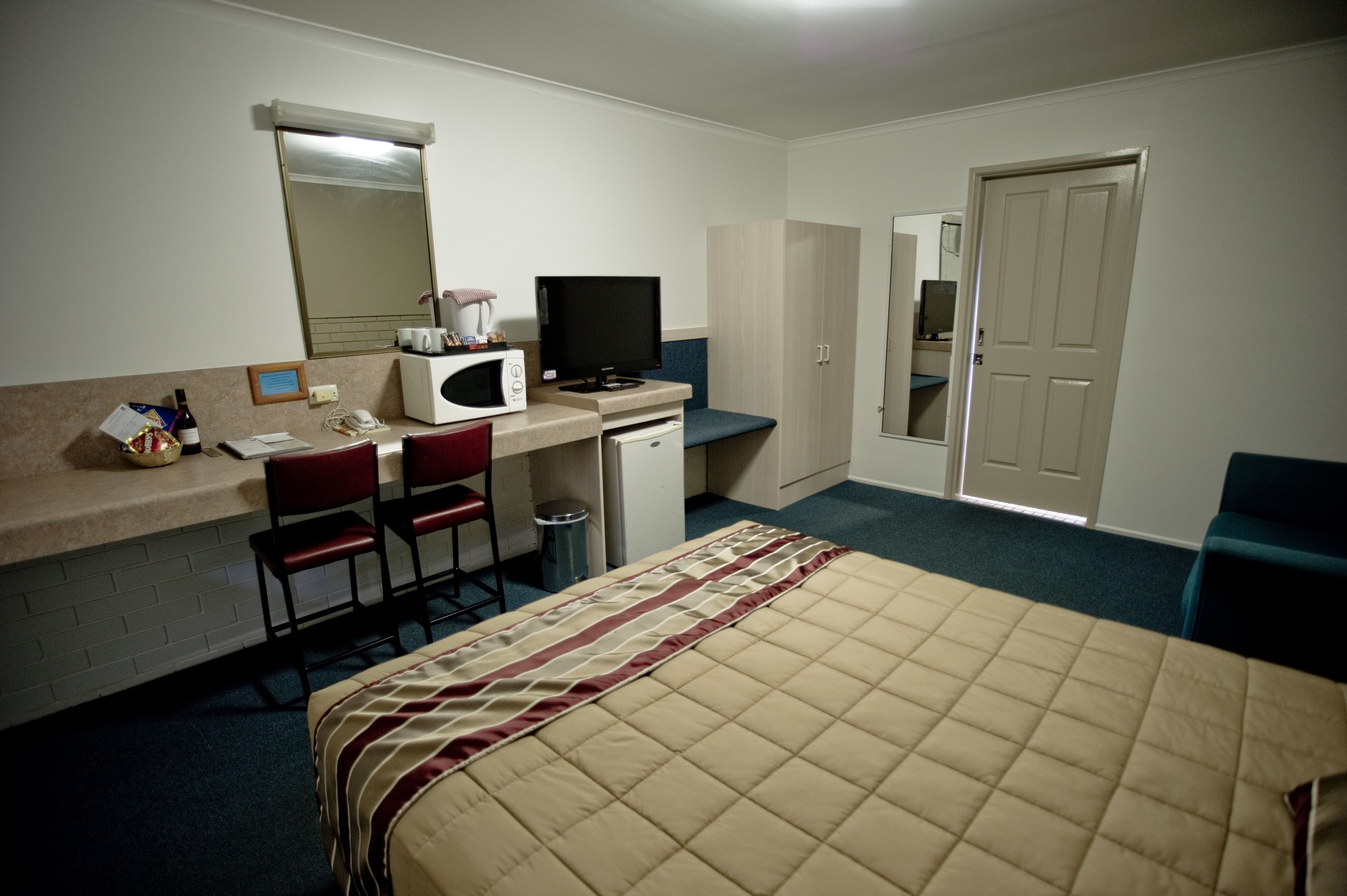 A hotel room with a bed, a desk with a computer and a TV, a chair, a microwave, a small trash bin, and a door.