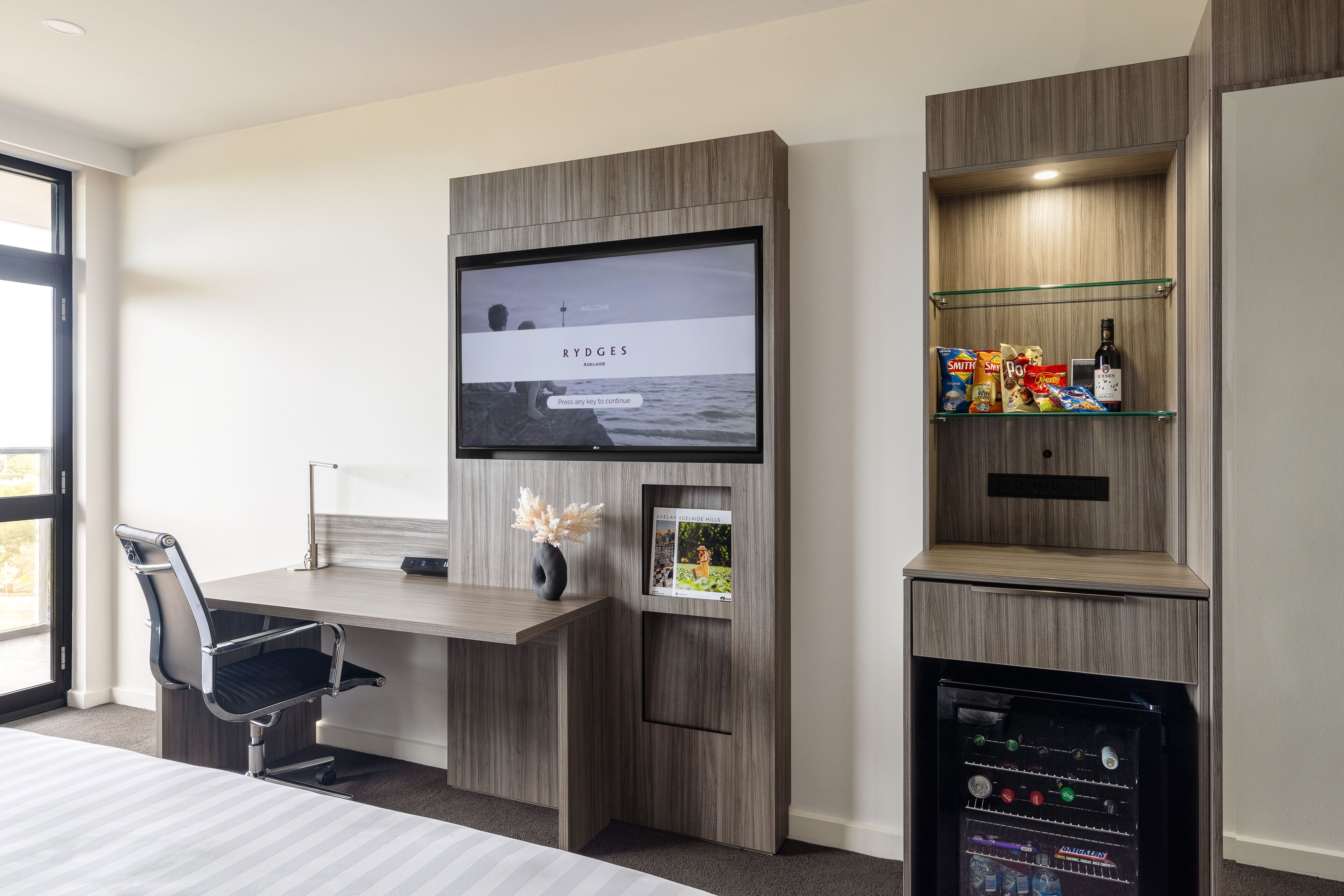 A modern hotel room with a wooden desk, a mounted TV, a minibar, and a glass shelf with snacks.