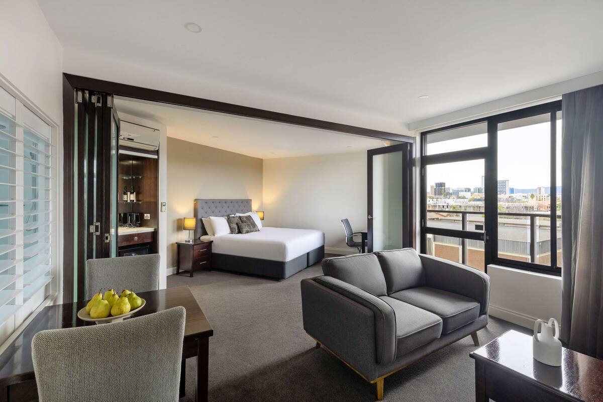 A hotel room with a bed, a sofa, a dining table with fruit, and a view of the city through large windows.